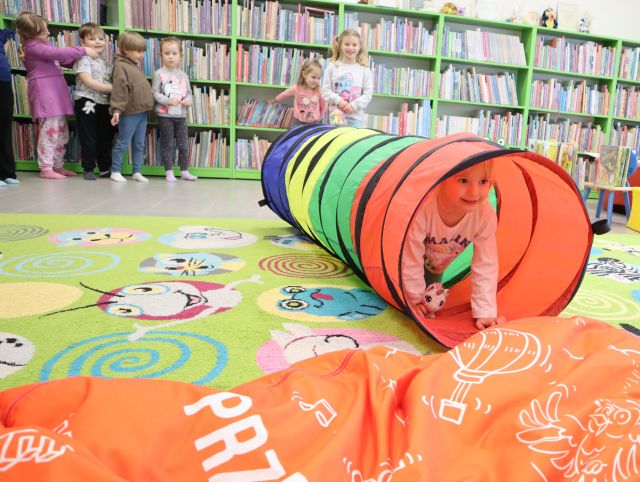 dzieci stoją przy regałach z książkami, dziewczynka wychodzi z tunelu sensorycznego