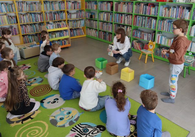 dzieci siedzą na kolorowym dywanie. Przed nmi siedzi kobieta z książką, w tle regały z książkami