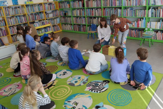 na kolorowym dywanie siedzą przedszkolaki, przed nimi kobieta i chłopiec, w tle regały z książkami