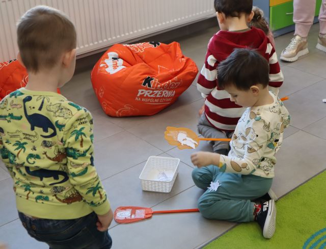 dzieci siedzą na podłodze, w rękach trzymają packi, w pudełęczkach leżą kartki