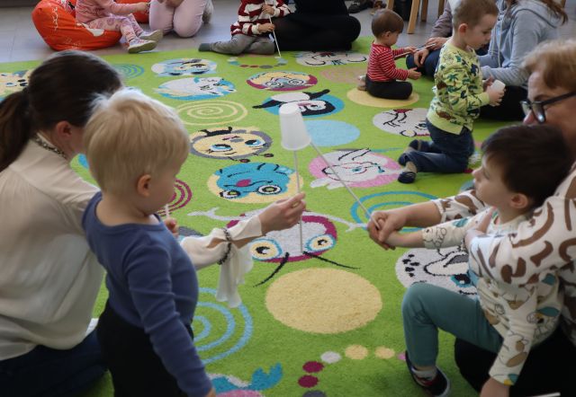 na kolorowym dywanie siedzą dizeci z mamami, mamy trzymają w dłoniach patyczki, na patyczkach plastikowe kubeczki