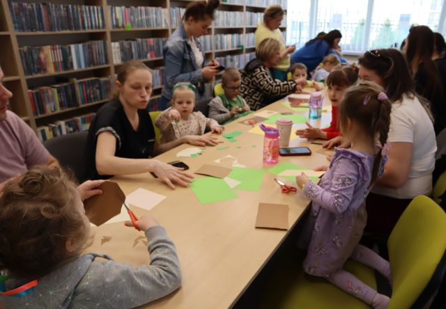 dzieci z rodzicami siedzą przy rzędzie stołów, na stołach materiały papiernicze