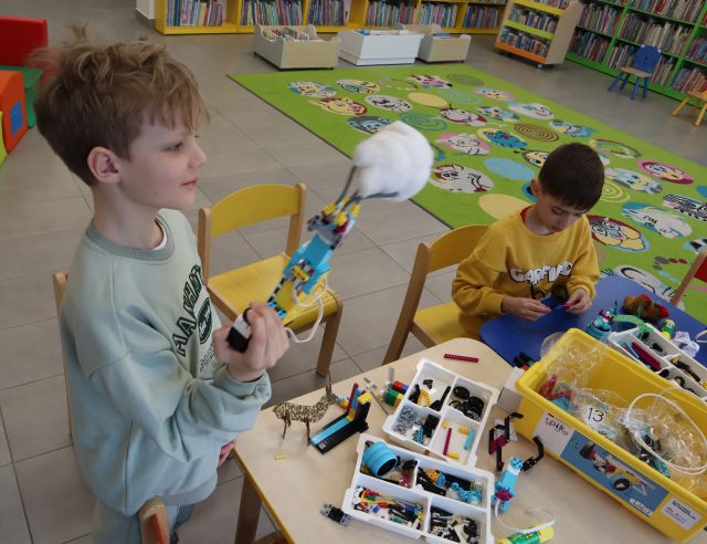 chłopiec z klockami LEGO, przy stole siedzi drugi chłopiec i buduje z klocków