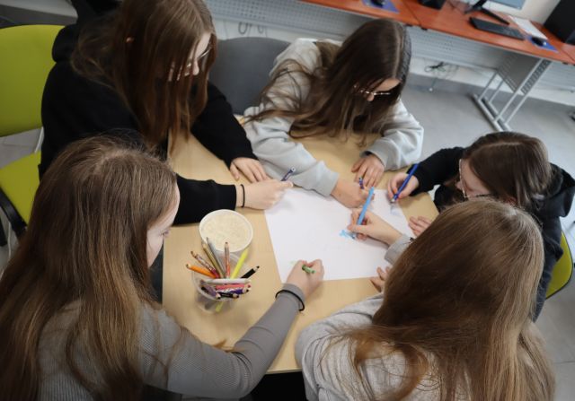 5 dziewczyn siedzi przy stole, na stole leży kartka, na której dziewczyny rysują