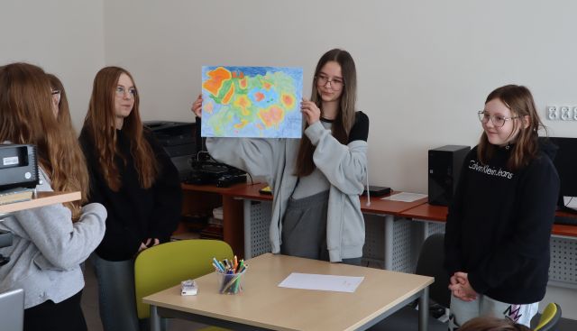dziewczyna prezentuje rysunek, obok stoją 3 dziewczyny, w tle stoliki z komputerami