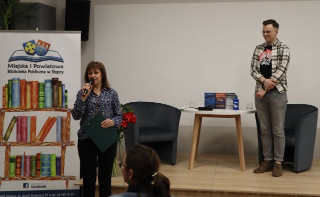 przed sceną stoi z mikrofonem dyrektorka biblioteki, na scenie mężczyzna, na scenie stolik i dwa fotele