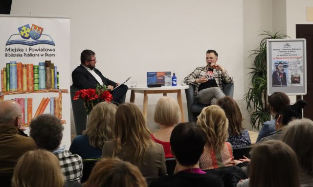 dwóch mężczyzn siedzi na fotelach na scenie, przed n imi siedzi publiczność