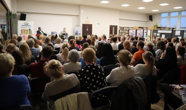 na zdjęciu publiczność siedząca na krzesłach, w tle na scenie siedzi dwóch mężczyzn