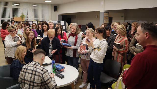 mężczyzna siedzi przy stoliku obok kobiety, przed nimi stoją uczestnicy spotkania