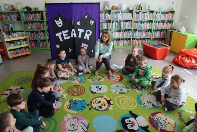 przedszkolaki siedzą na dywanie, przed nimi na krzesełku siedzi bibliotekarka, w tle tablica z napisem teatr oraz regały z książkami