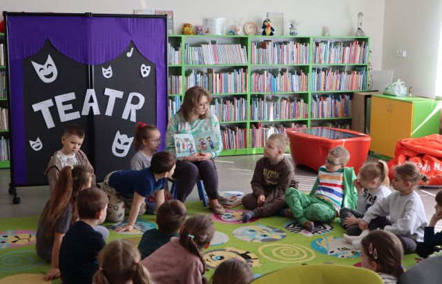 na dywanie siedzą przedszkolaki, przed nimi na krzesełku siedzi bibliotekarka z książką, w tle tablica z napisem teatr oraz regały z książkami
