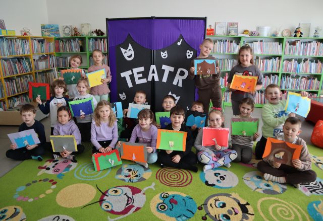 przedszkolaki pozują do zdjęcia z wykonanymi pracami, w tle regały z książkami, oraz tablica z napisem teatr