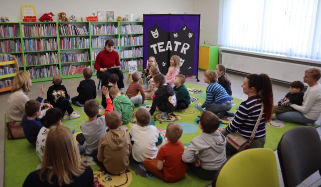 przedszkoalki siedzą na kolorowym dywanie, przed nimi siedzi na krzesełku bibliotekarka, w tle regały z książkami