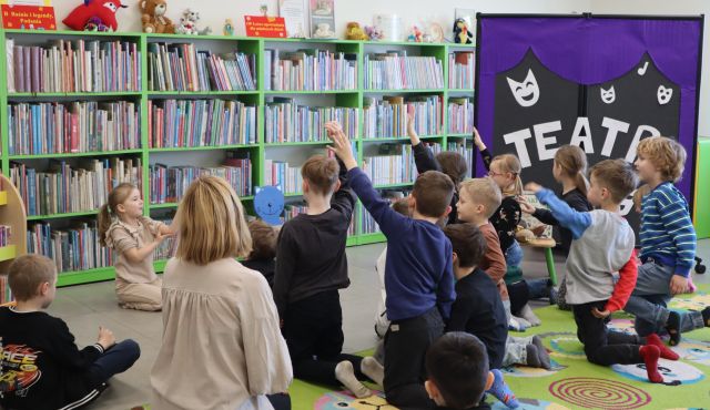 na kolorowym dywanie siedzą i klęczą przedszkolaki z podniesionymi rękami, w tle regały z książkami