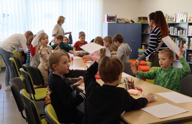 przedszkolaki siedzą na krzesełkach przy stołach i rysują na kartkach
