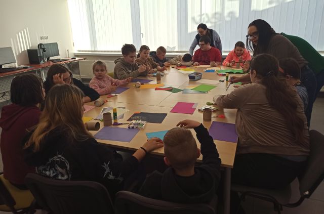 młodzież siedzi przy połączonych stołach, na których leżą kolorowe kartki i inne materiały papiernicze