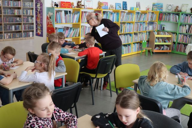 dzieci siedzą przy stolikach, w tle regay z książkami