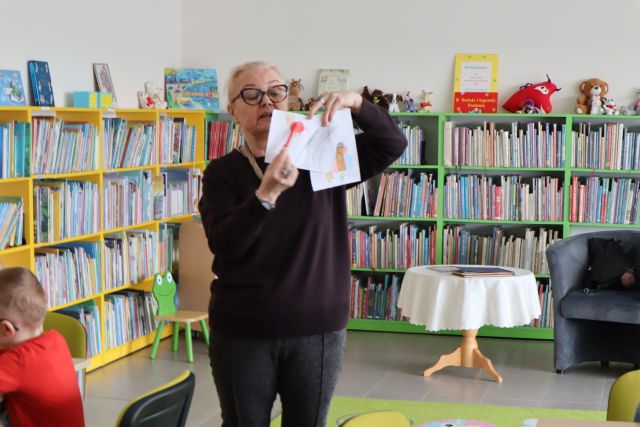 kobieta pokazuje kartki z rysunkami, w tle regały z książkami