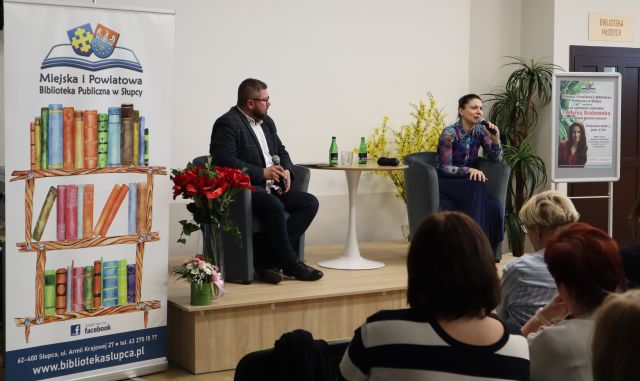 na scenie przy stole na fotelach siedzi mężczyzna i kobieta przed publicznością