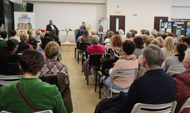 na zdjęciutyłem siedząca na krzesłąch publiczność, w tle mężczyzna i kobieta siedzą na scenie