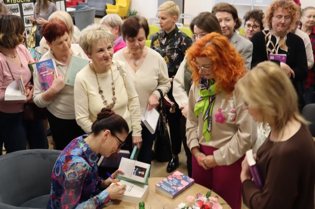 kobieta siedzi przy stoliku i podpisuje książkę, wokól niej stoją uczestniczki spotkania