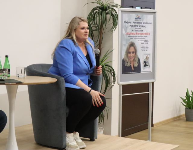 kobieta siedzi przy stoliku, obok plakat z zaproszeniem na spotkanie