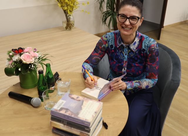 autorka z długopisem w dłoniach uśmiecha się do aparatu, na stole leżą książki