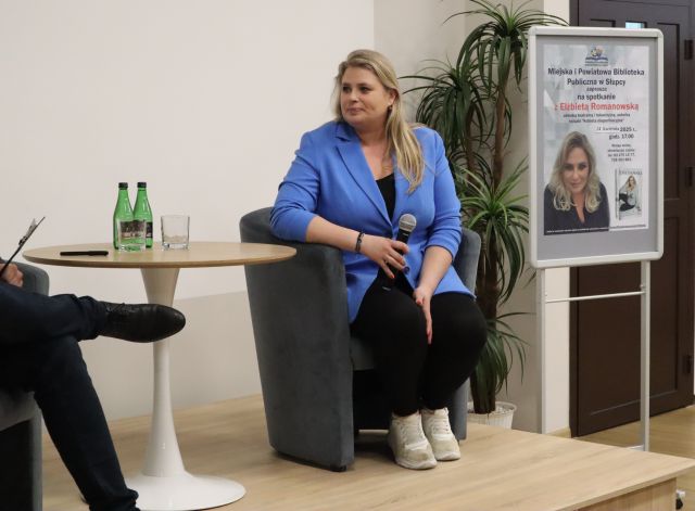 kobieta siedzi przy stoliku, obok plakat z zaproszeniem na spotkanie