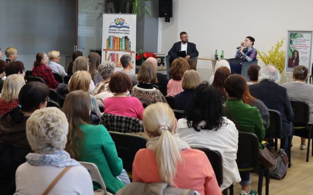 na scenie przy stole na fotelach siedzi mężczyzna i kobieta przed publicznością