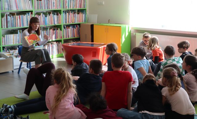 dzieci siedzą na kolorowym dywanie, przed nimi na krześle siedzi bibliotekarka z książką, w tle regały z książkami