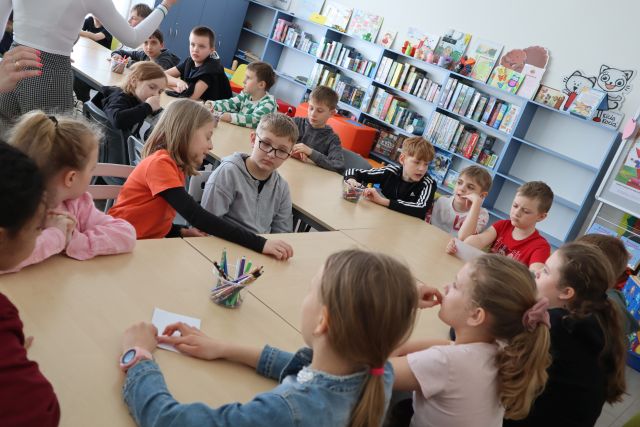 dzieci siedzą przy stołach ustawionych w podkowę, w tle regały z książkami