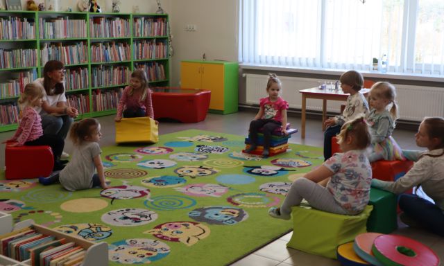 dzieci i bibliotekarka z książką siedzą na kolorowym dywanie i pufach