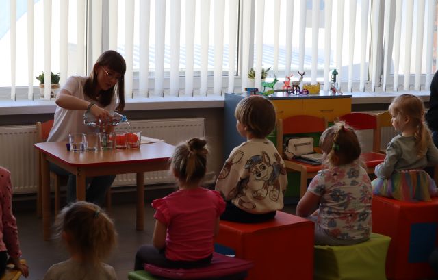 bibliotekarka robi przy stoliku eksperyment z kolorowaniem  papierków, przed nią na pufach siedzą dzieci