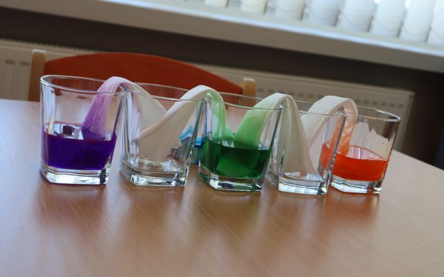 na stole 3 szklanki z kolorowymi papierkami i wodą