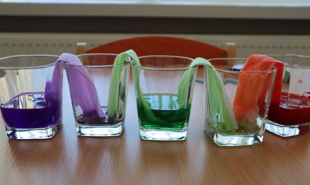 na stole 3 szklanki z kolorowymi papierkami i wodą