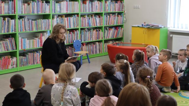 bibliotekarka z książką siedzi na krzesełku przed przedszkolakami siedzącymi na dywanie, w tle regały z książkami