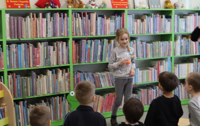 dziewczynka z piłeczką stoi przed kolegami z przedszkola, w tle regały z książkami