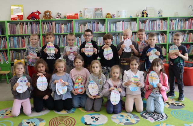 do zdjęcia pozują przedszkolaki ze swoimi pracami przedstawiającymi kolorowe pisanki, piierwszy rząd siedzi na dywanie, drugi rząd siedzi, w tle regały z książkami