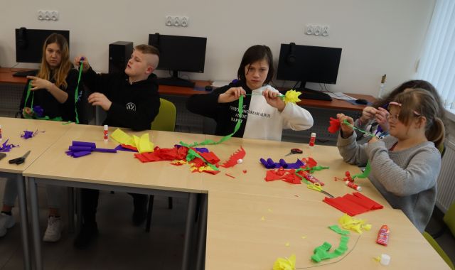 młodzież siedzi przy stołach, na stołach bibuła i nożyczki, w tle komputery