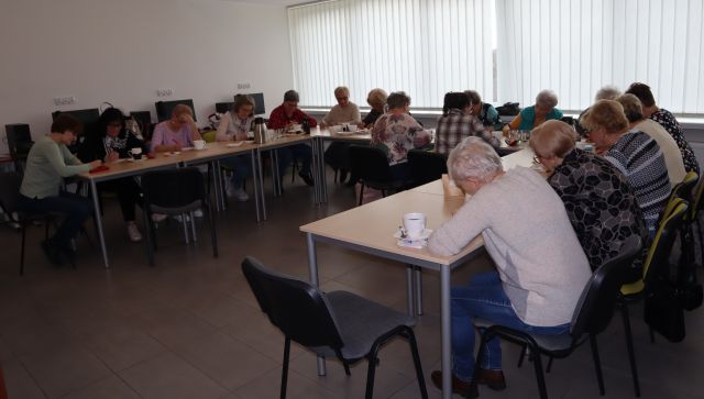grupa około 20 seniorek siedzi przy stołavch pochylona nad kartkami