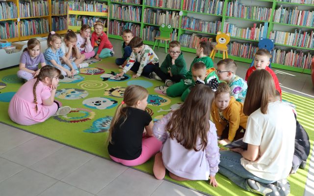 na kolorowym dywanie siedzą przedszkolaki, w  tle regały z książkami