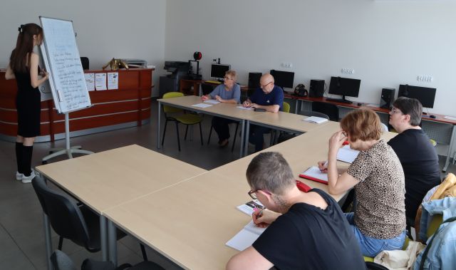 przy stolikach siedzą seniorzy i notują, przed nimi dziewczyna przy flipcharcie