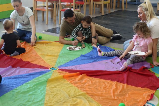 rodzice z dziećmi siedzą na chuście animacyjnej,a na chuście leżą materiały plastyczne