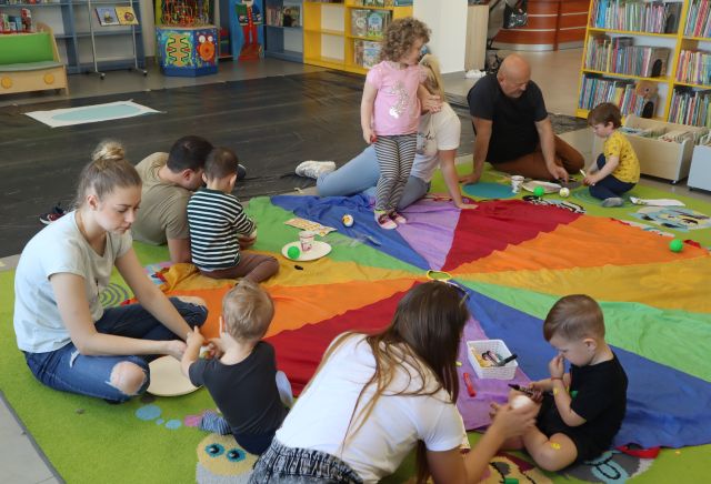 dzieci z rodzicami siedzą na chuście animacyjnej, na chuście leżą materiały plastyczne