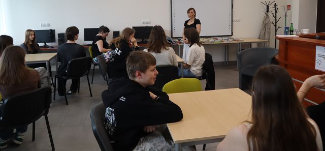 młodzież siedzi przy stolikach, przed nimi stoi kobieta, w tle biurka z komputerami