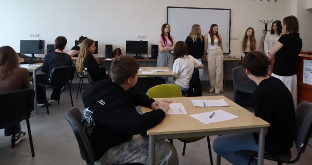 część młodzieży siedzi przy stolikach, przed nimi stoi grupka młodzieży