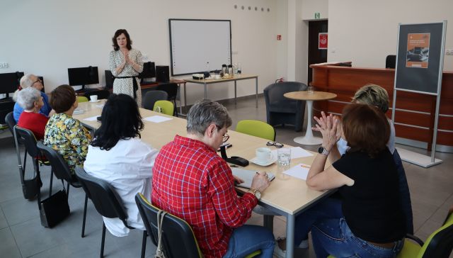 przy stolikach siedzą seniorki pochylone na kartkami, przed nimi stoi kobieta, w tle komputery