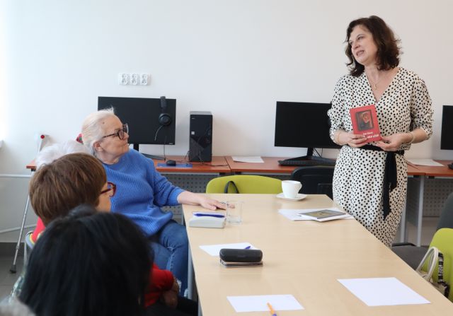 przy stolikach siedzą seniorki, przed nimi stoi kobieta, w tle komputery