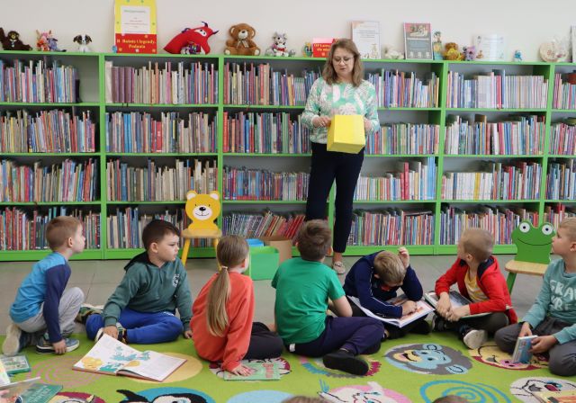 przedszkolaki isedzą na kolorowym dywanie, przed nimi stoi bibliotekarka, w tle regały z książkami