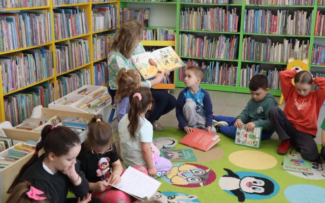przedszkolaki siedzą na kolorowym dywanie, przed nimi na krzesełku siedzi z książką bibliotekarka, w tle regały z książkami